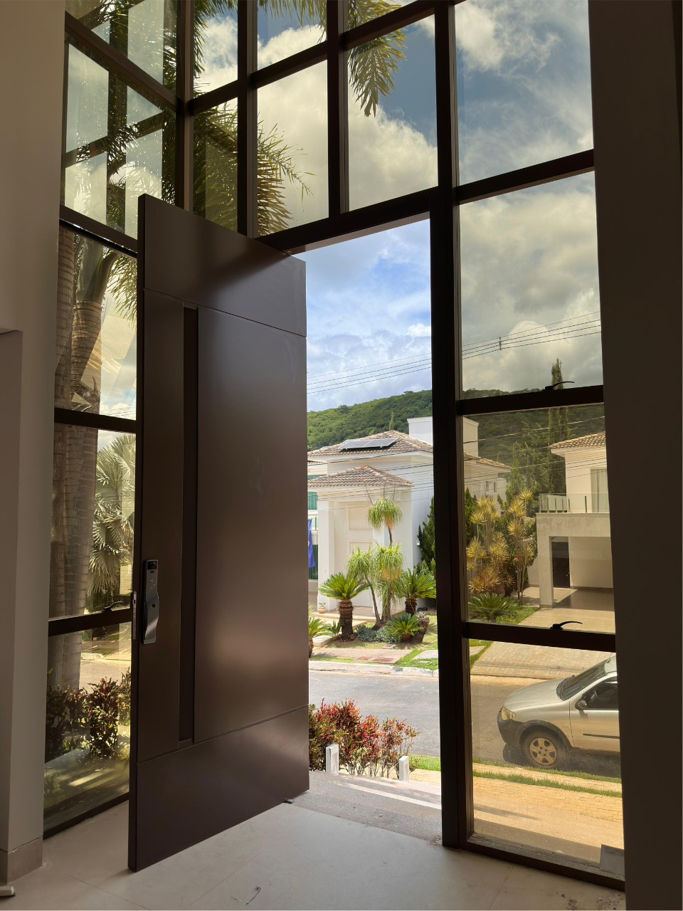 Entrada residencial com porta pivotante em ACM marrom integrada a painéis de vidro fixos.